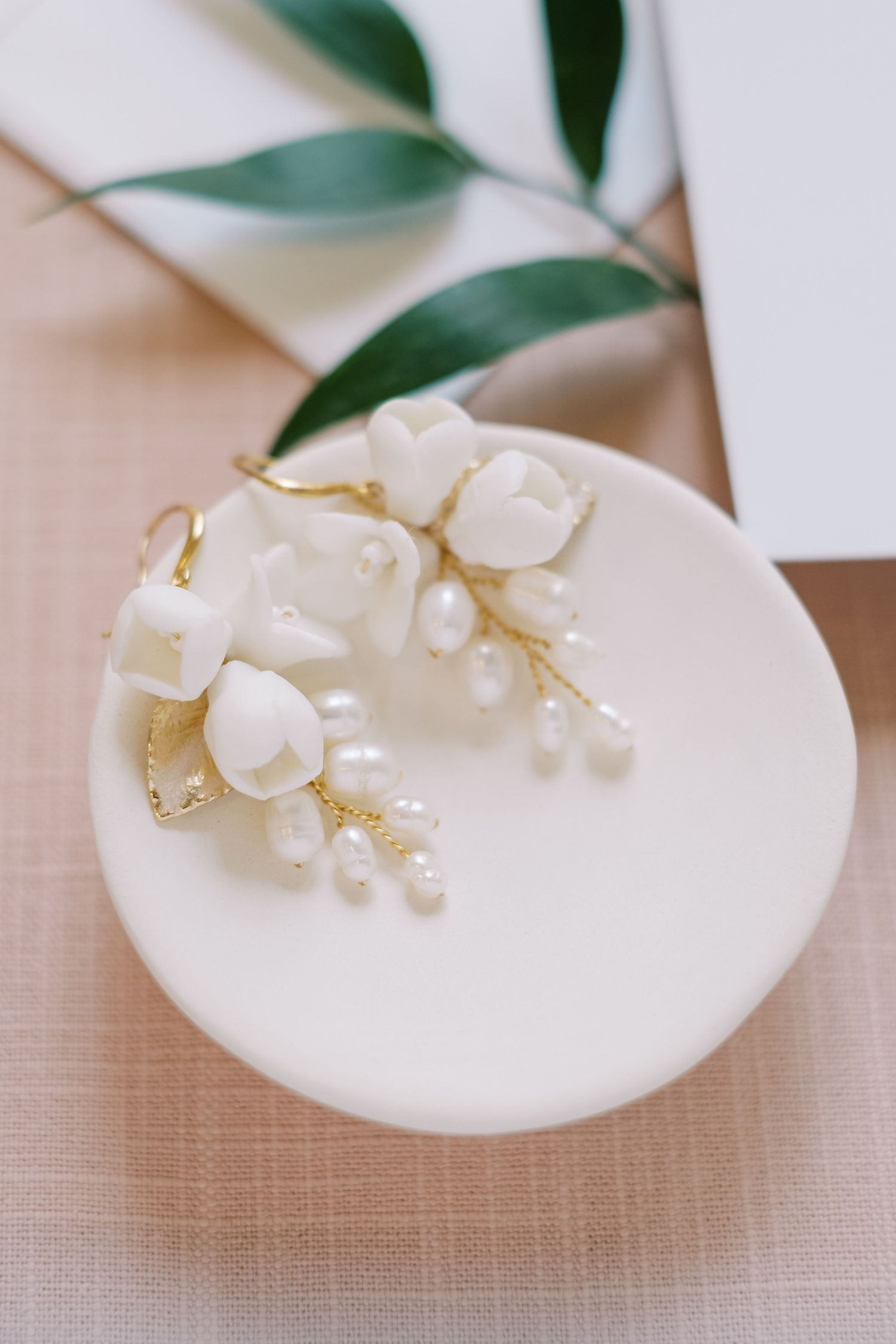 Pearl and gold bridal earrings displayed on white ceramic dish with greenery.