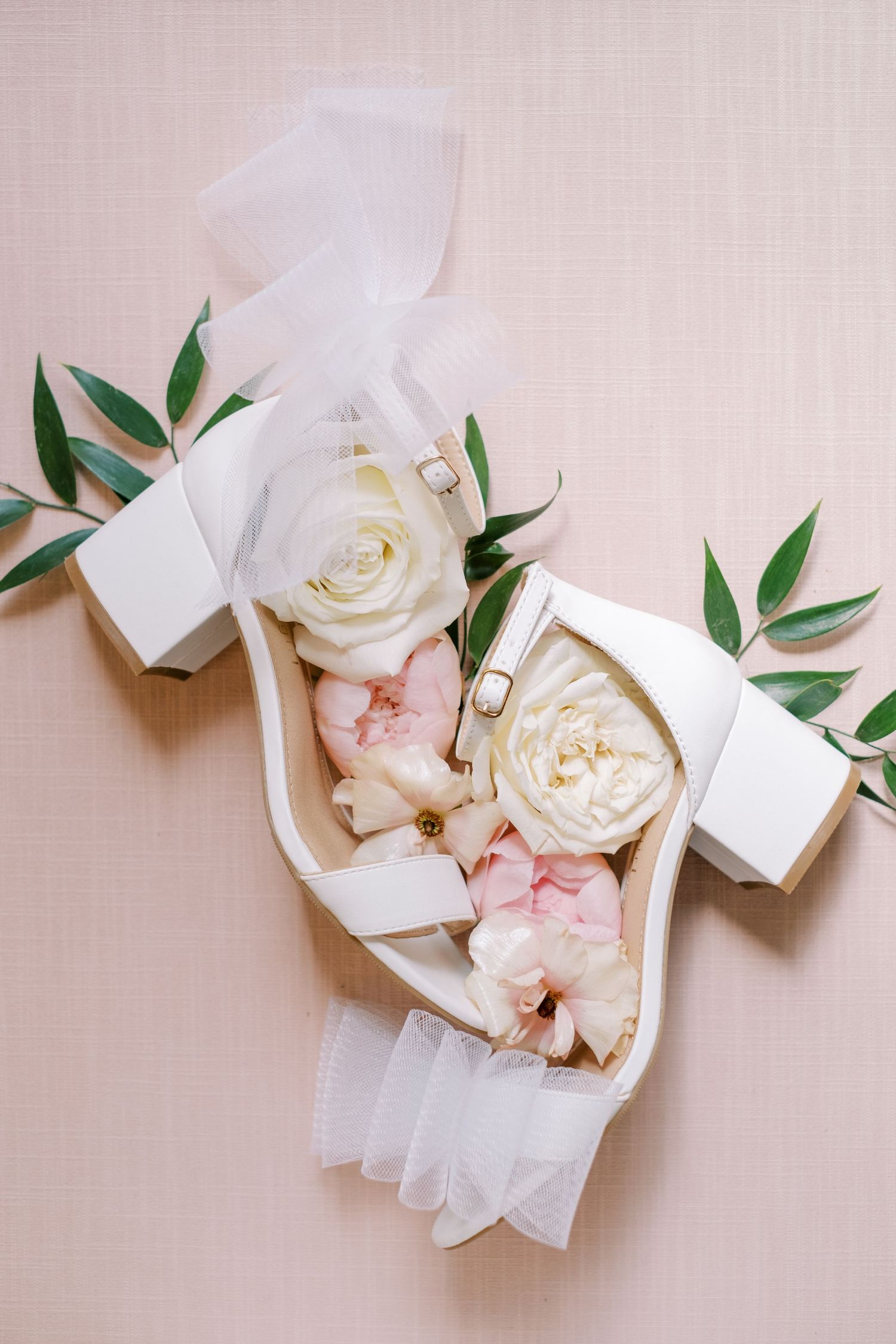 Elegant white bridal shoes decorated with delicate roses and greenery against a neutral background.
