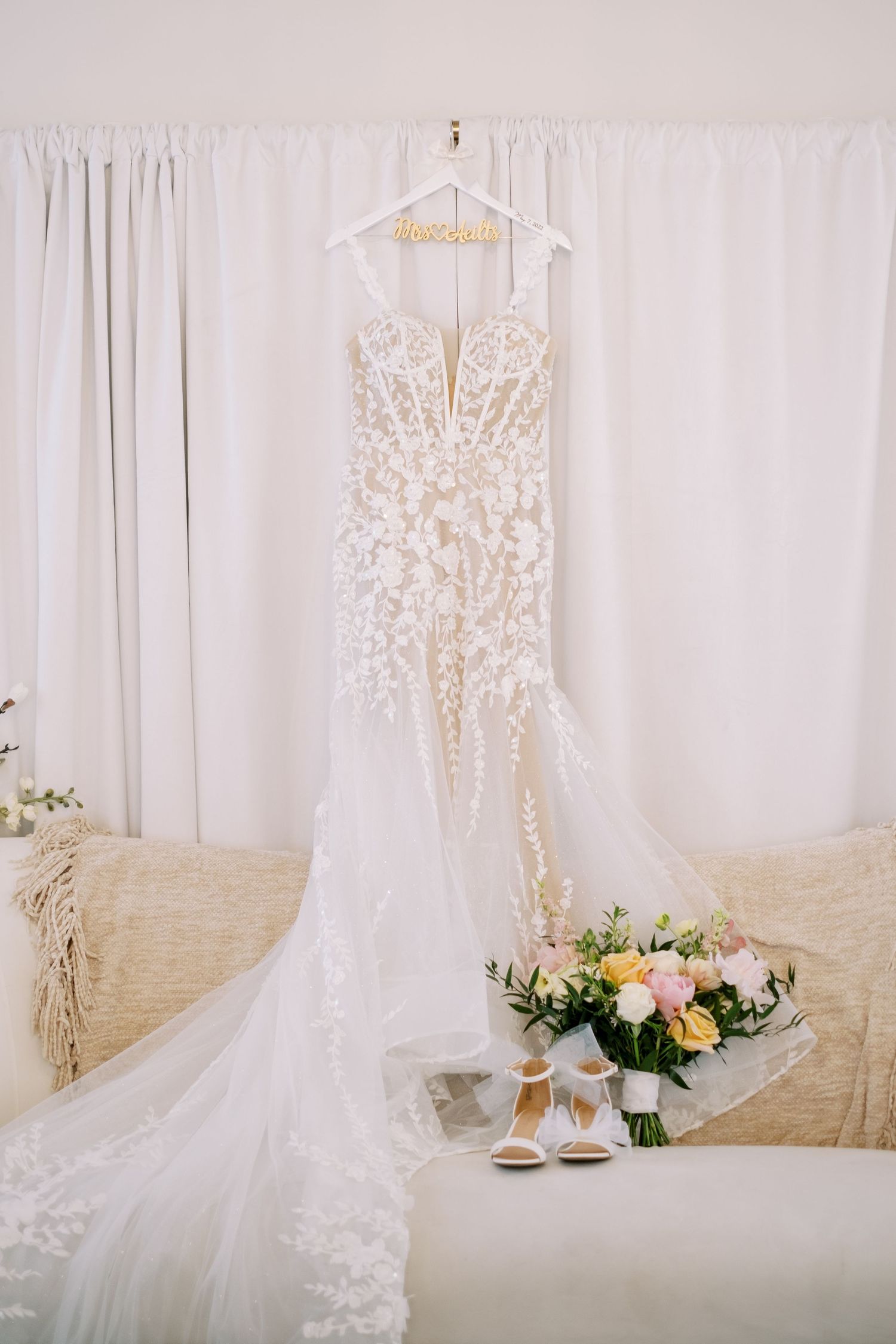 Ethereal white lace wedding dress hanging against soft white curtain backdrop.