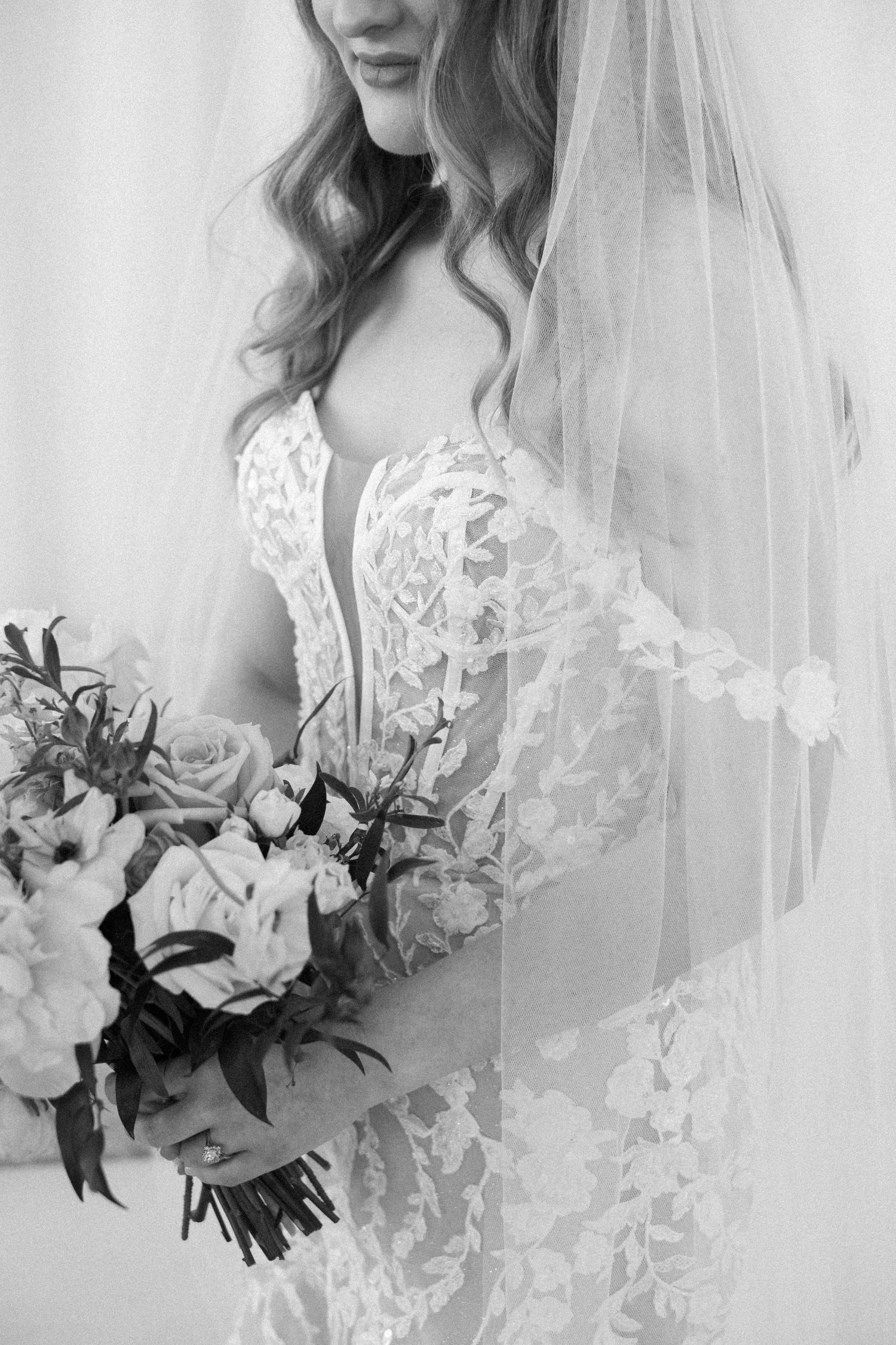 Delicate lace wedding dress with flowing veil and white floral bouquet captured in black and white.