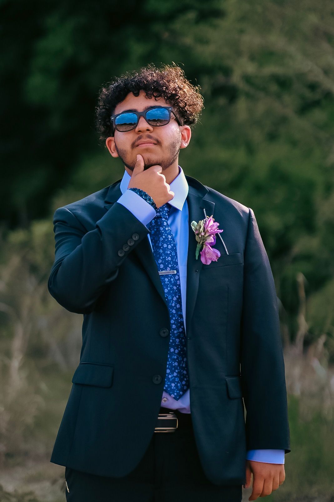 Young man wearing suit and tie and sunglasses holds his chin while looking at the camera