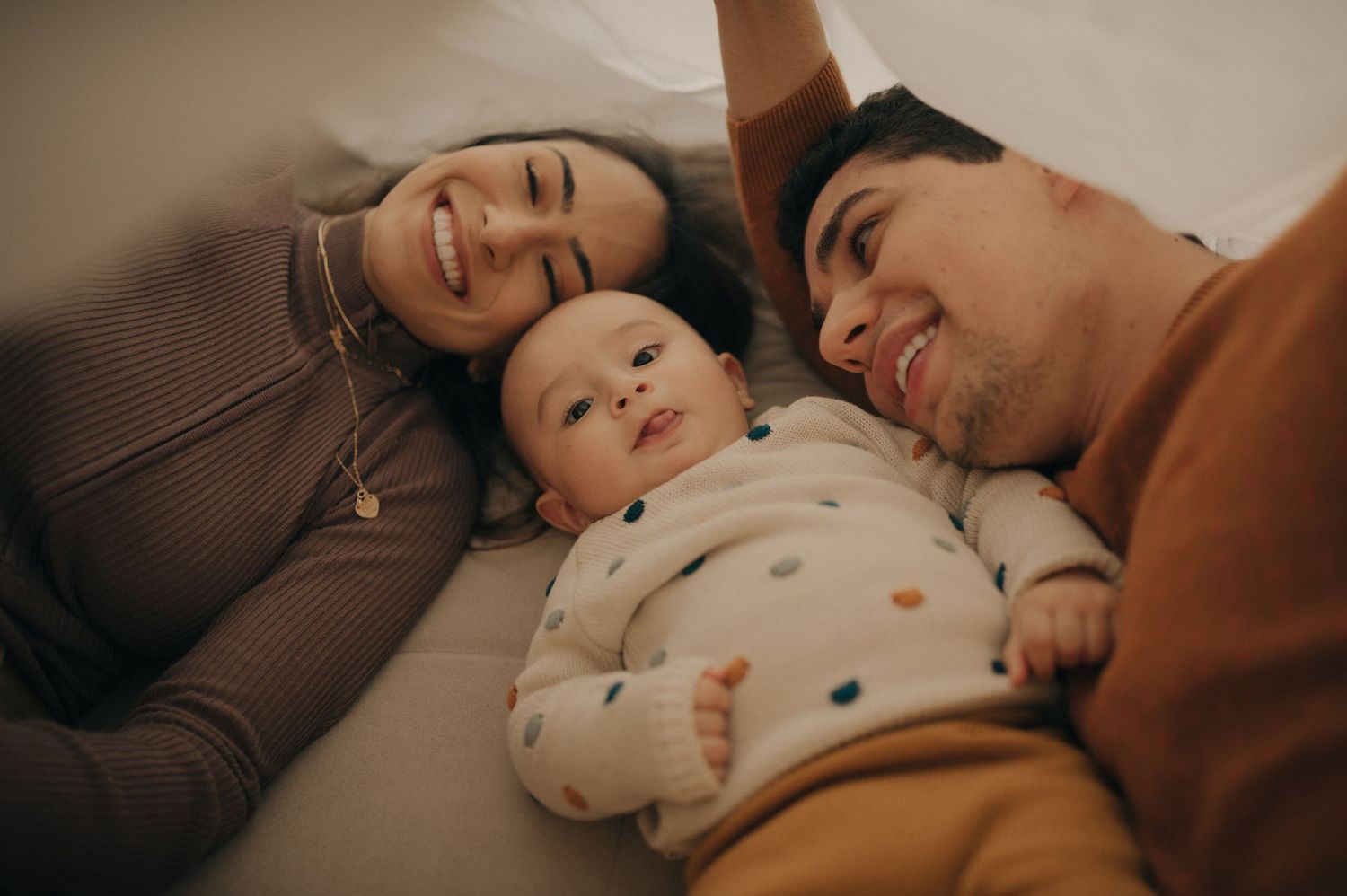 Family lying together on couch sharing tender moment with baby in polka dot outfit.
