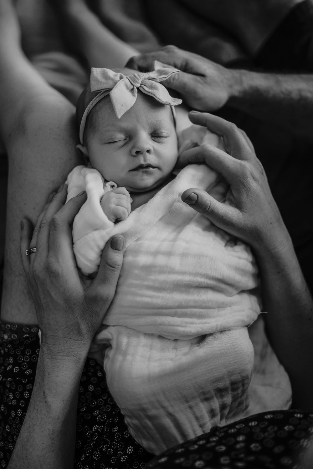 A sleeping newborn baby wearing a bow headband is gently cradled by caring hands.