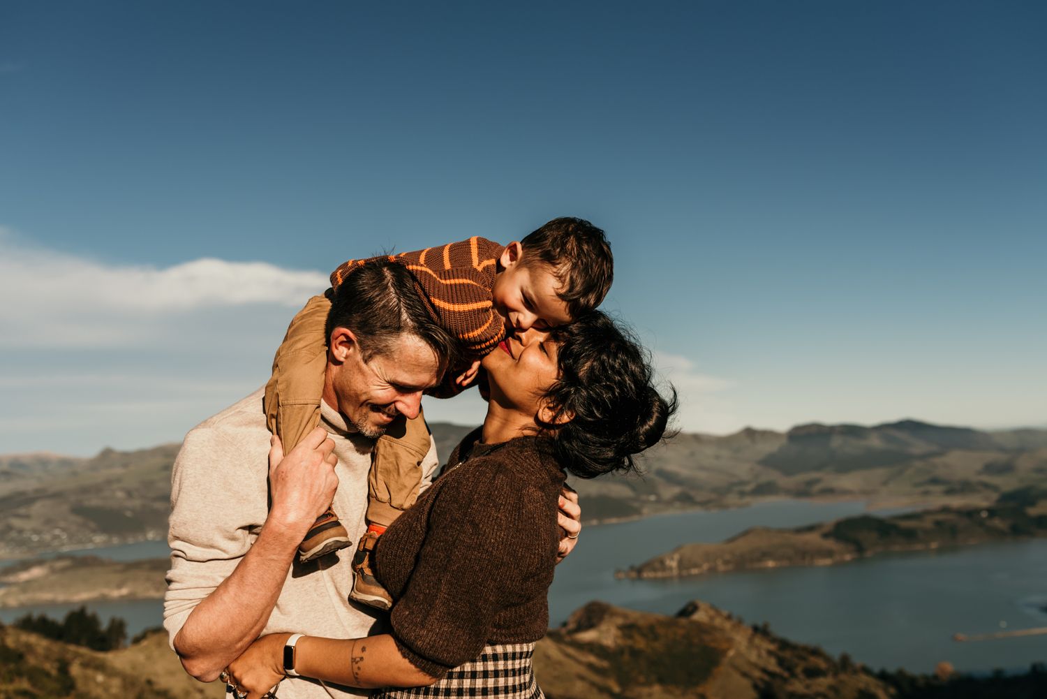 Tender family moment captured against dramatic mountain landscape with lake view at sunset.
