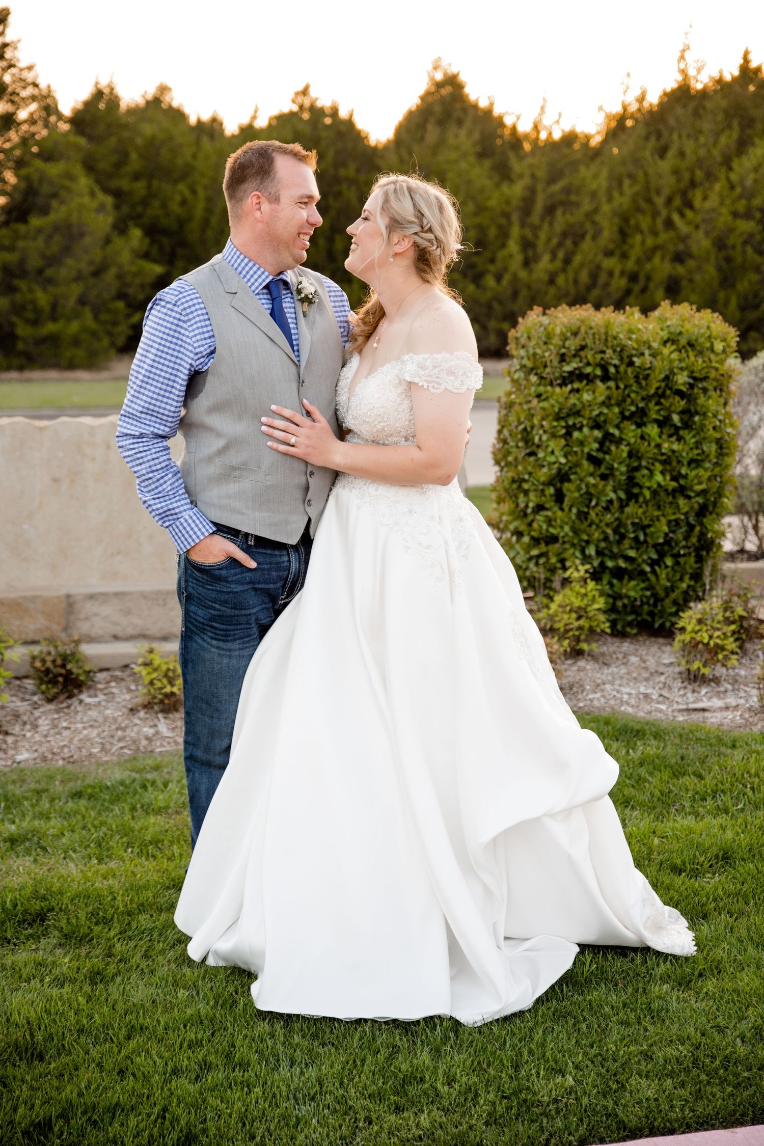 Wedding couple embracing outdoors with bride in flowing white dress and groom in vest and jeans.