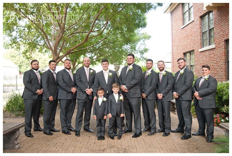 Groomsmen in dark suits lined up against brick building exterior.