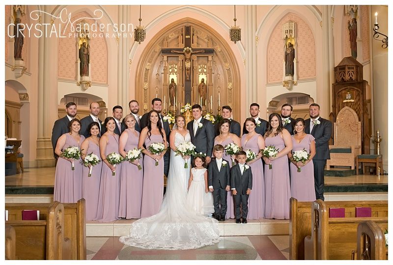 Wedding party in purple bridesmaid dresses and black tuxedos posed inside ornate church.