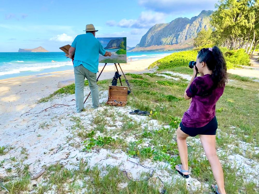 Artist paints a seascape on an easel while being photographed at a scenic Hawaiian beach with mountains in background.