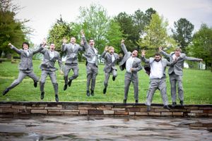 Wedding party in matching gray suits jumps joyfully on a brick ledge in an outdoor garden setting.