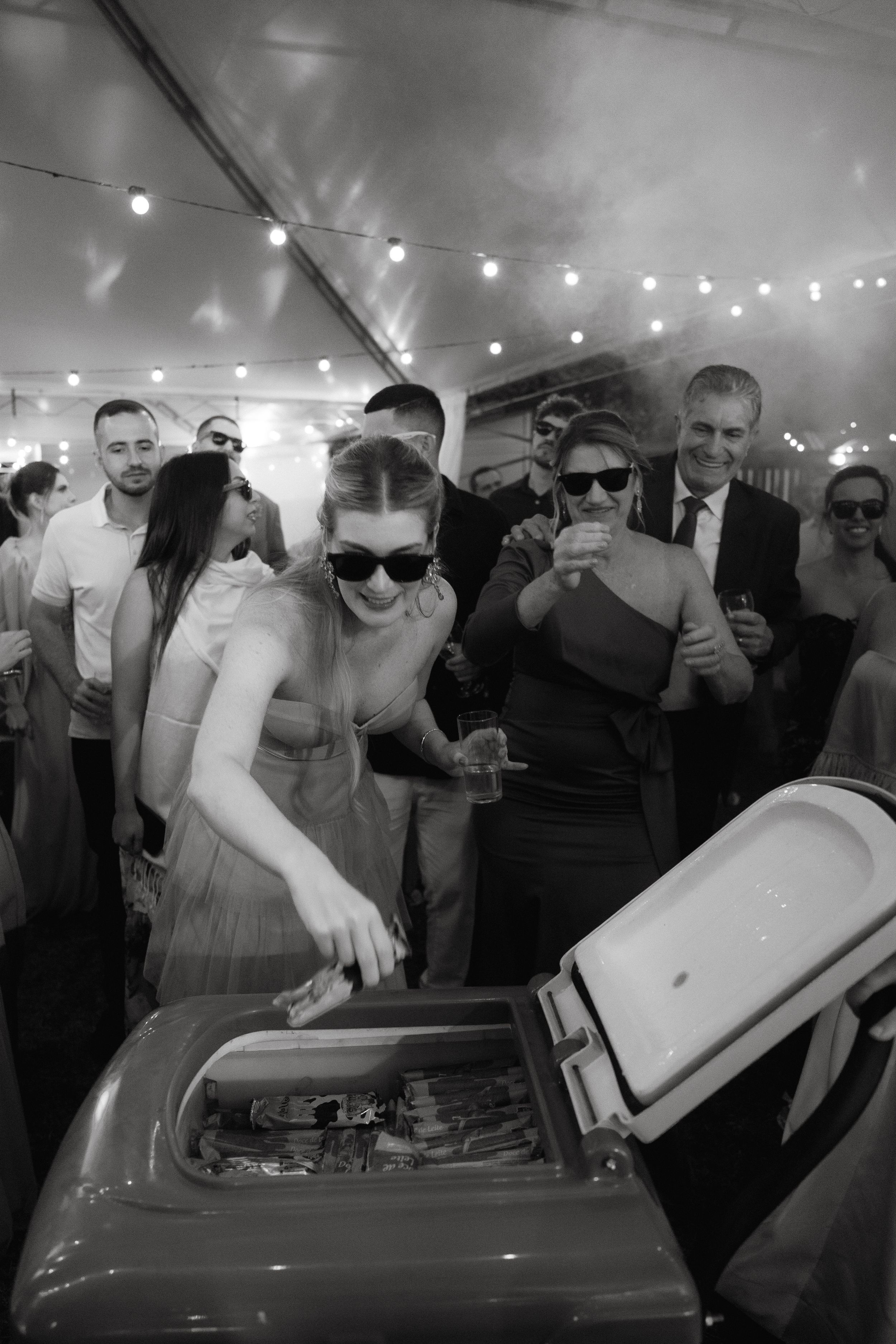 Wedding guests in sunglasses gather around cooler during outdoor reception under string lights.