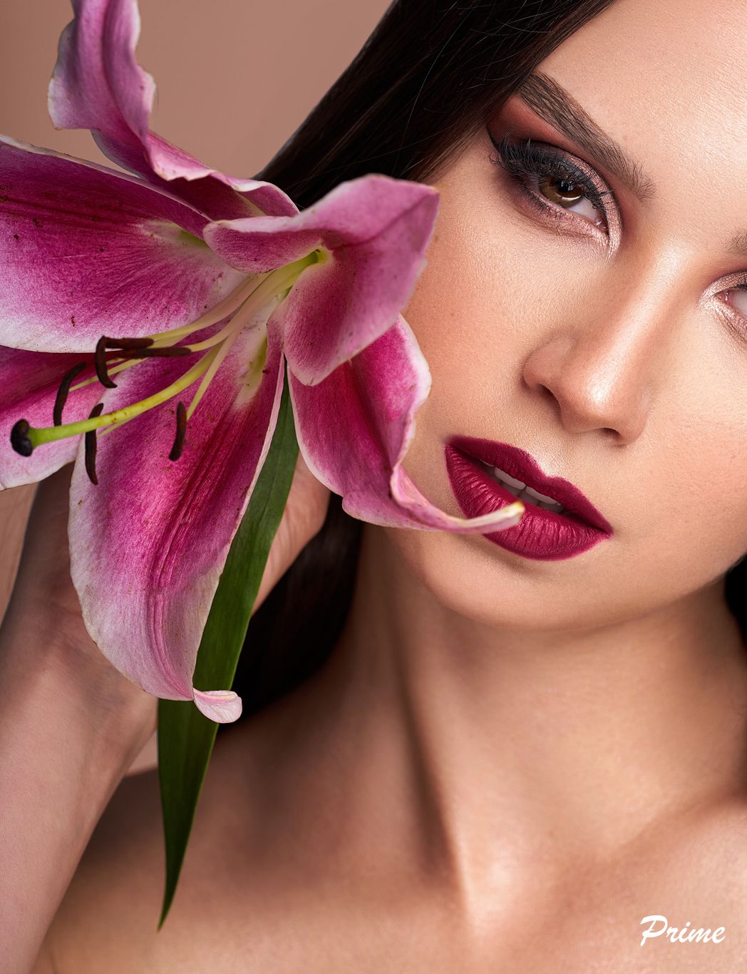 A vibrant pink stargazer lily bloom positioned next to crimson lips and dramatic eye makeup in a beauty portrait.
