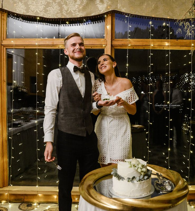 A couple stands together by their wedding cake surrounded by twinkling fairy lights.