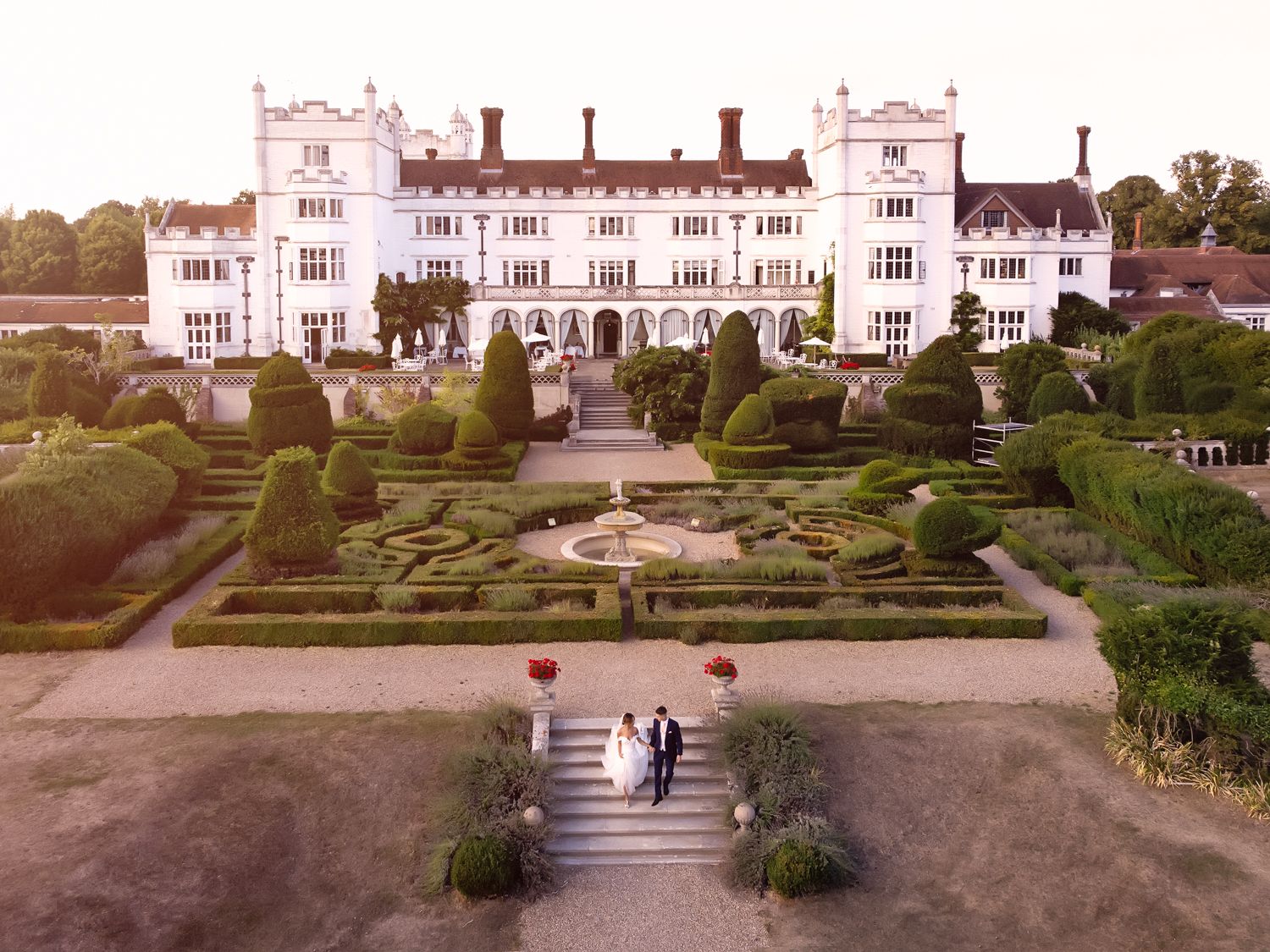Aerial view of newlyweds walking through formal gardens towards grand manor house.