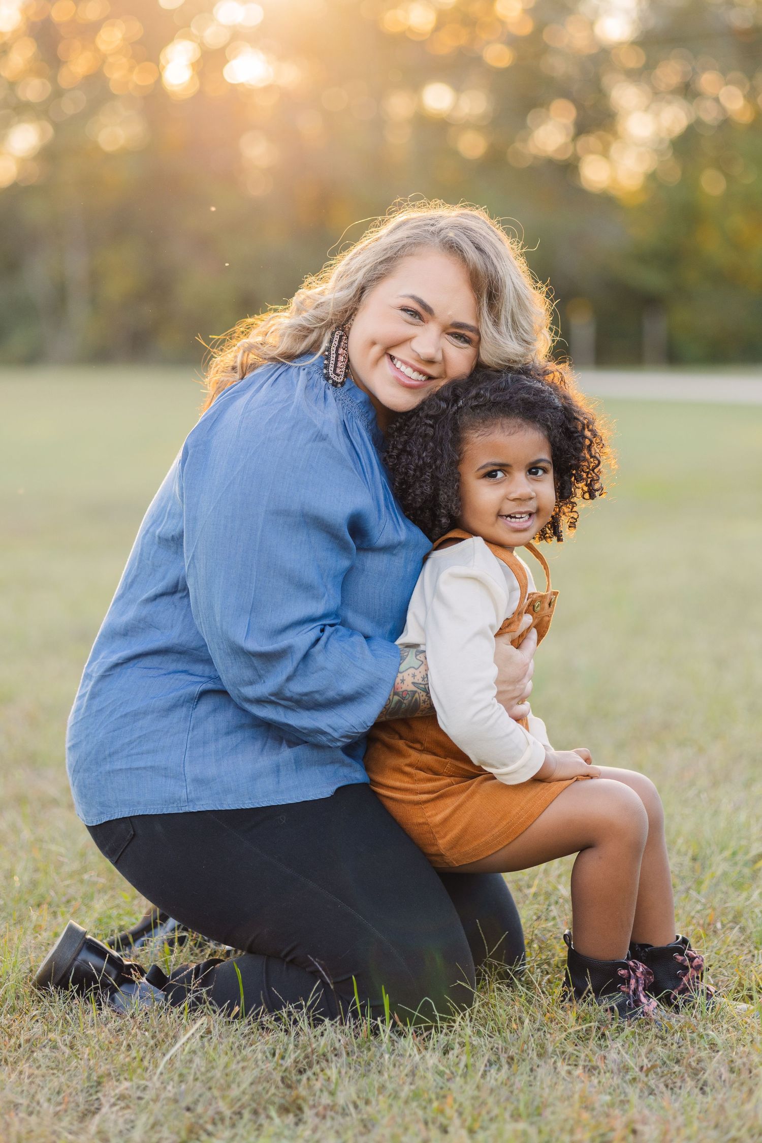 mother in a blue shirt holds daughter in field in an orange outfit during golden hour