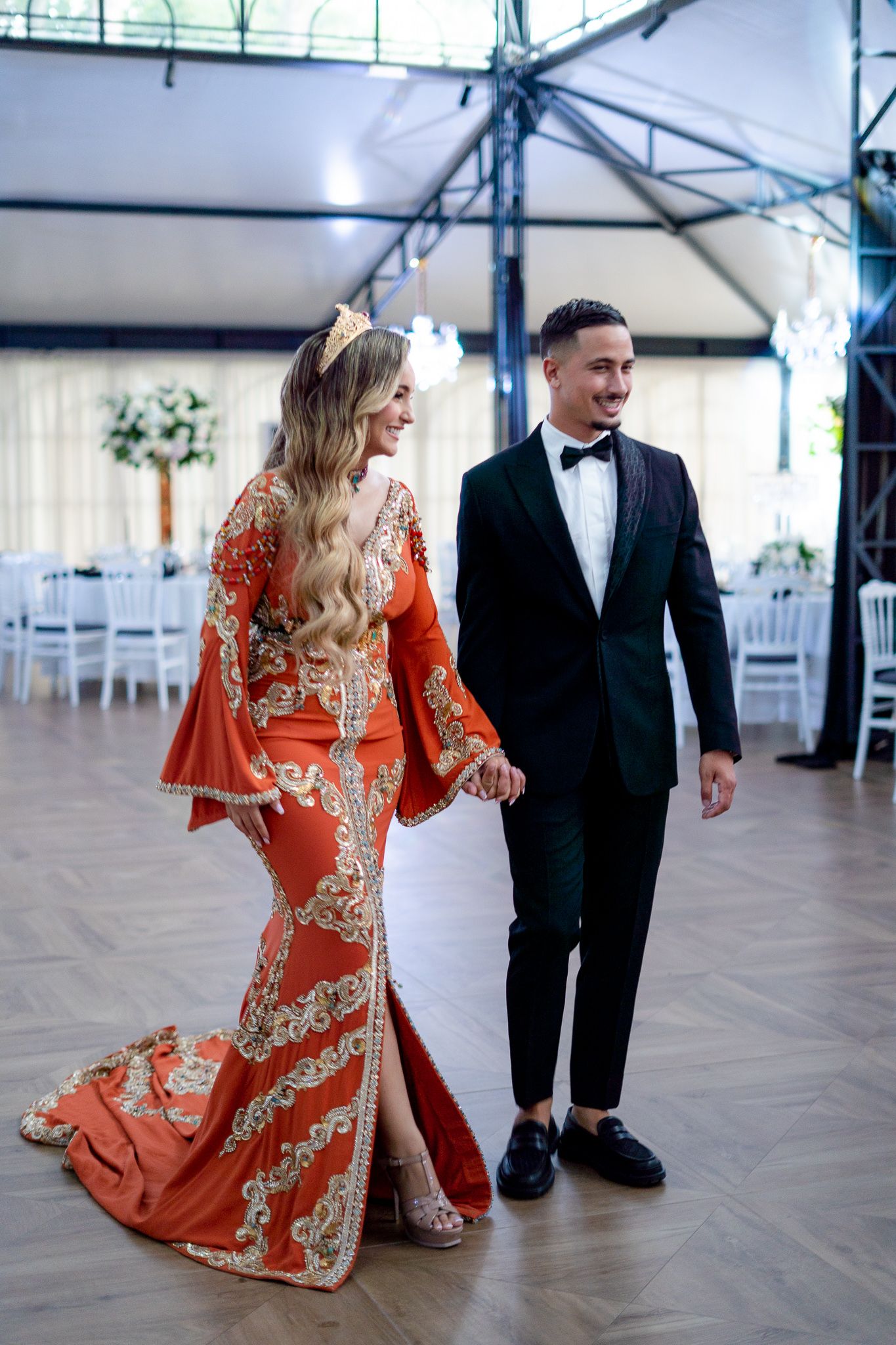 A couple in formal attire, with a woman in an orange patterned dress and man in black tuxedo, walking together indoors.