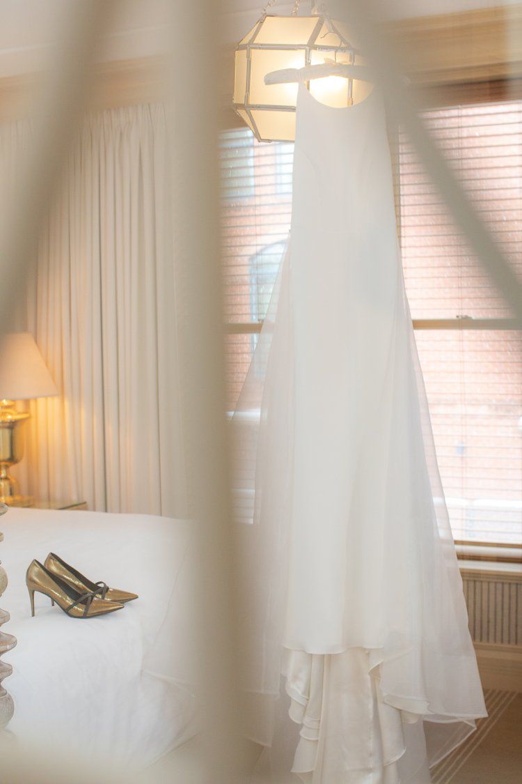White wedding dress hanging by window with soft natural lighting and heels in foreground.