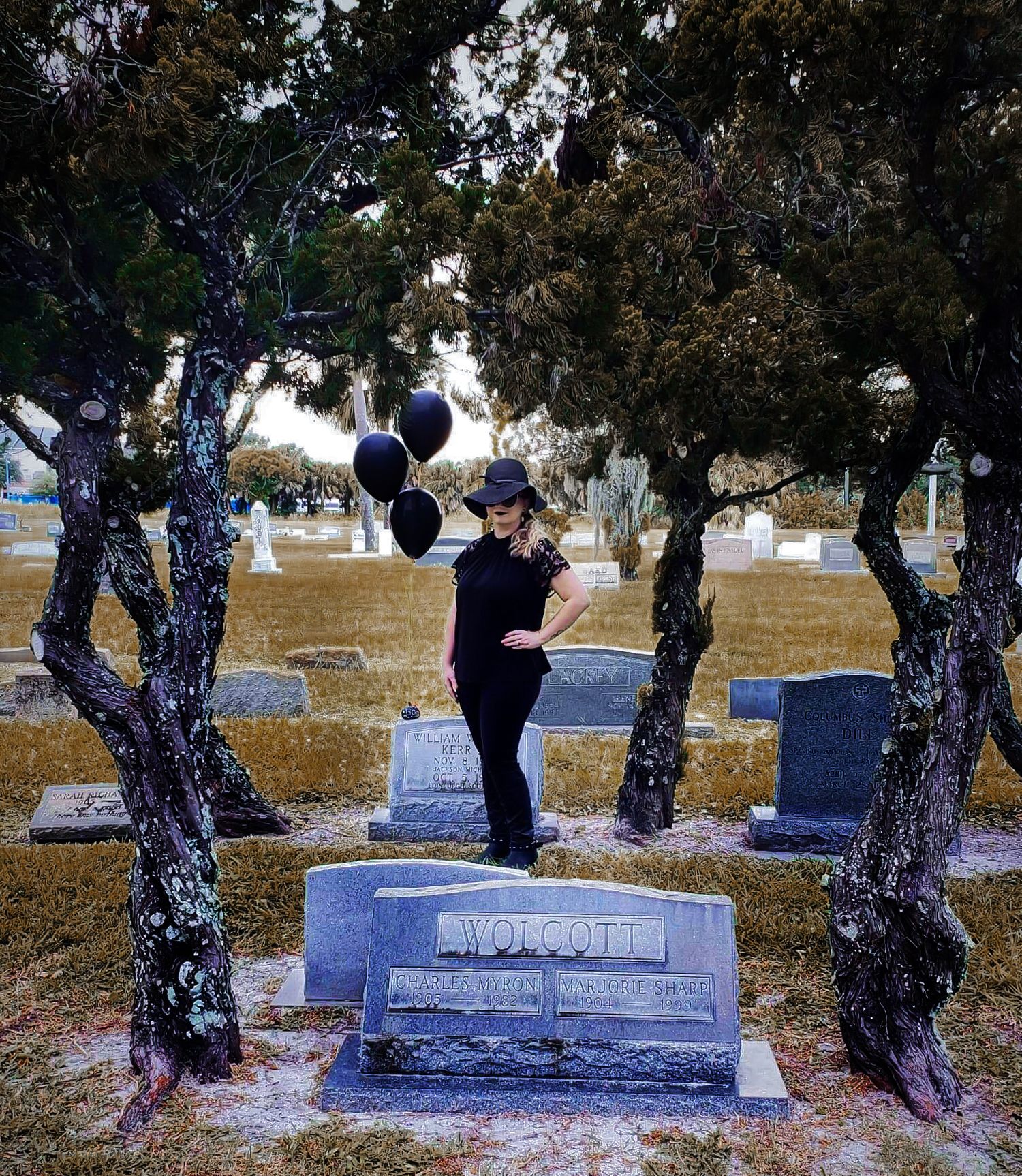 Woman in black outfit holding dark balloons standing among gravestones in a scenic cemetery.