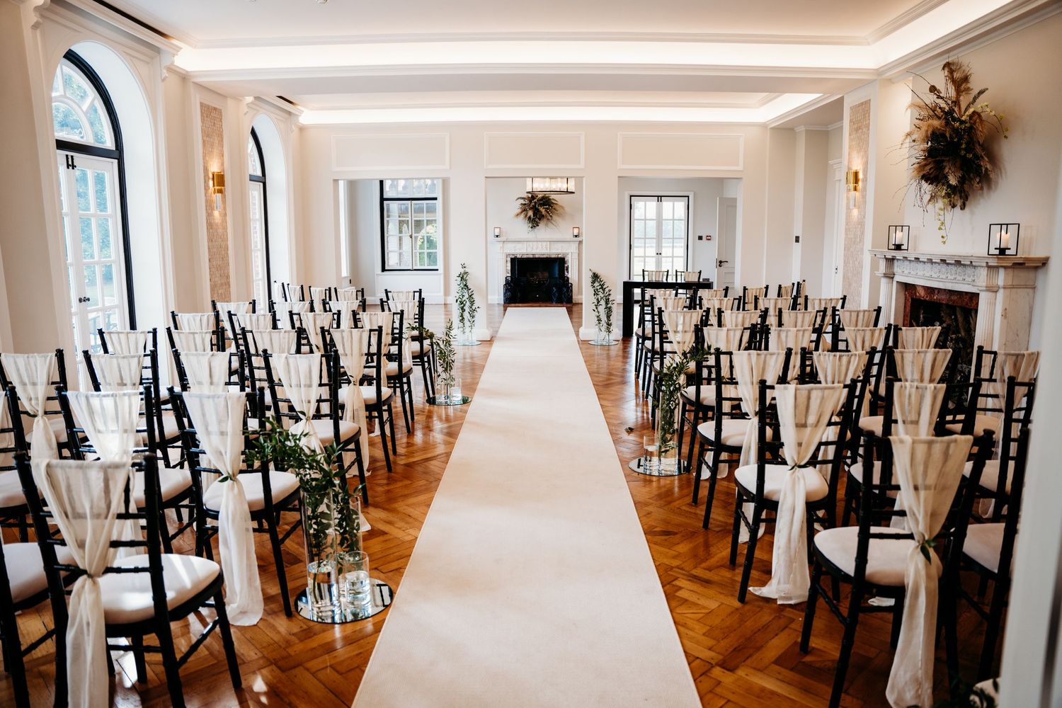 Elegant wedding ceremony setup with black chairs, white aisle runner, and floral decor in a bright indoor venue.
