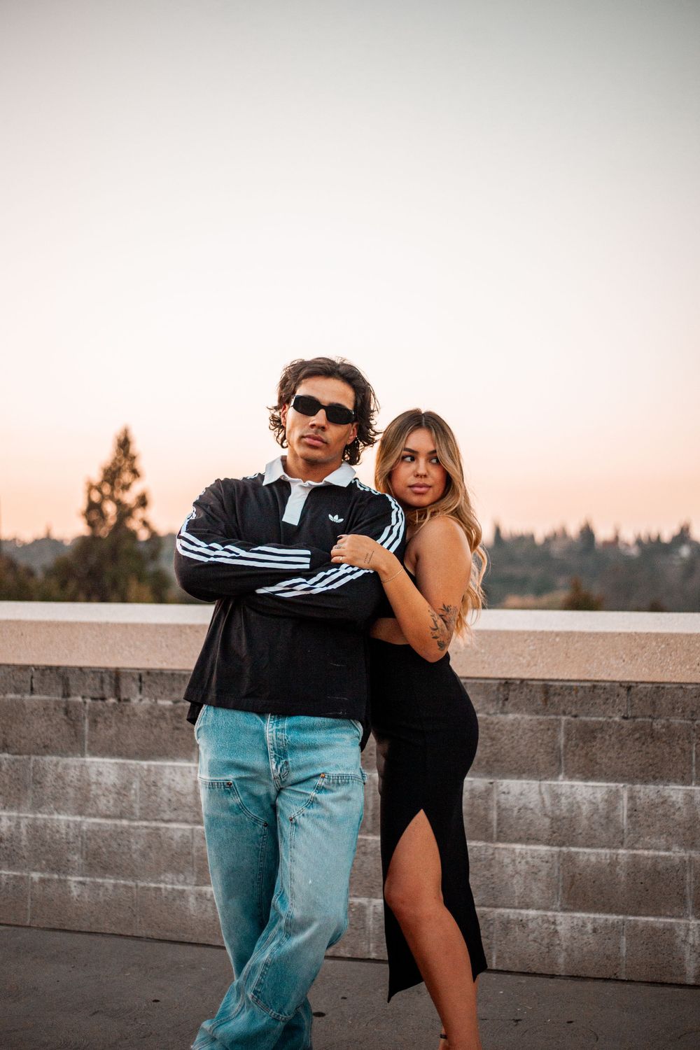 Couple posing together against concrete wall during sunset, wearing casual attire.