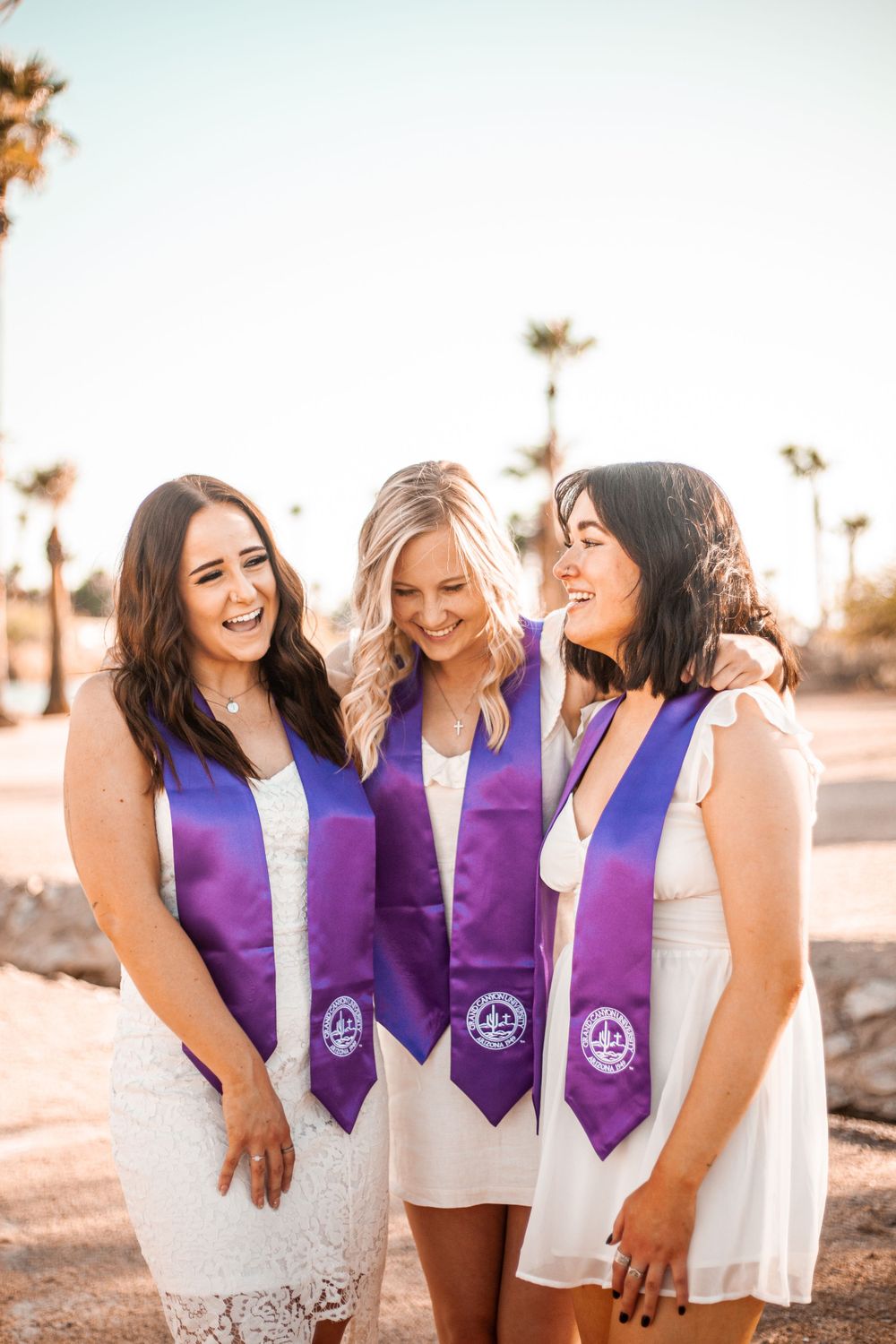 Three friends in white dresses wearing matching purple graduation stoles celebrating together outdoors.