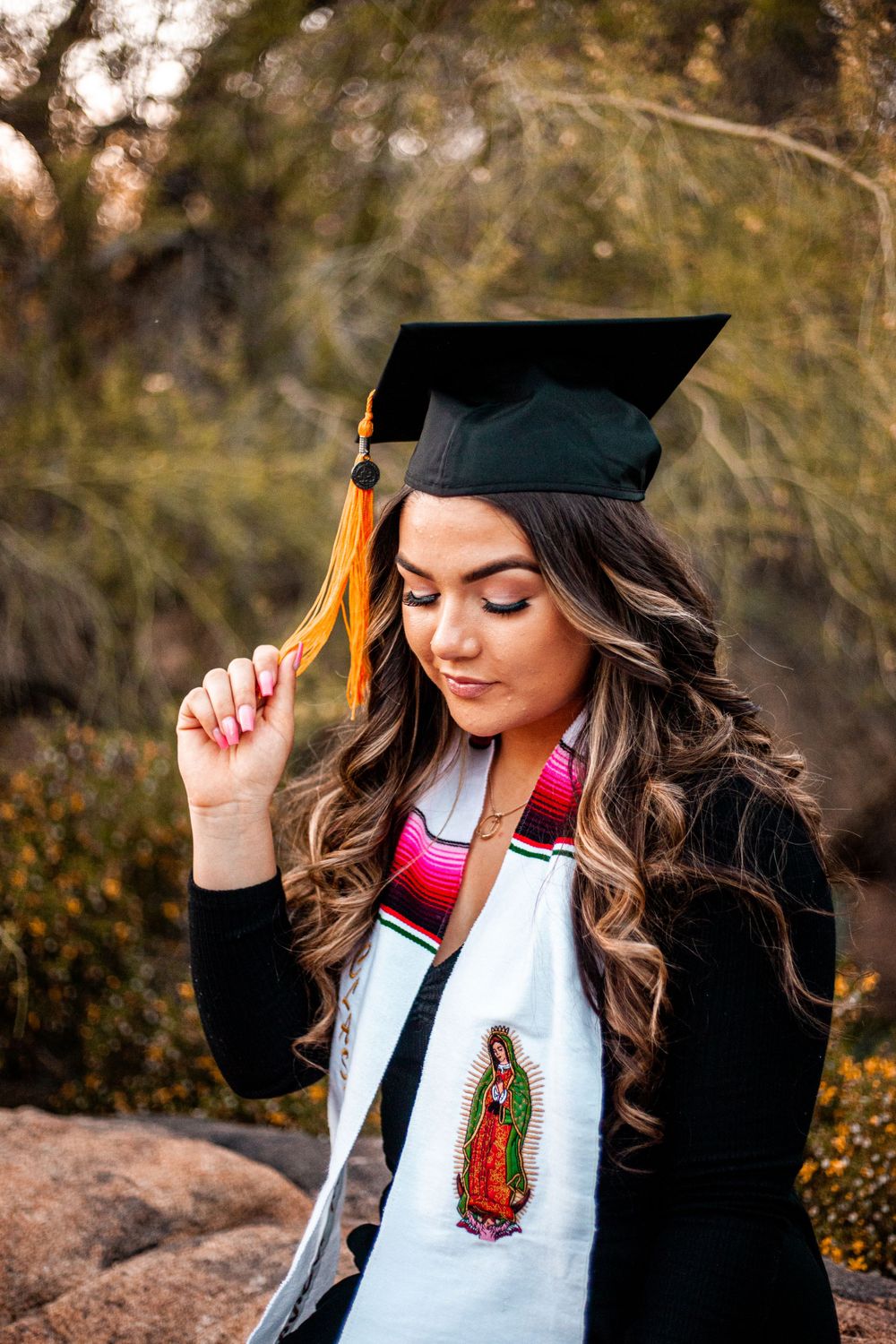 Graduate in cap and gown poses thoughtfully with religious imagery on graduation stole.
