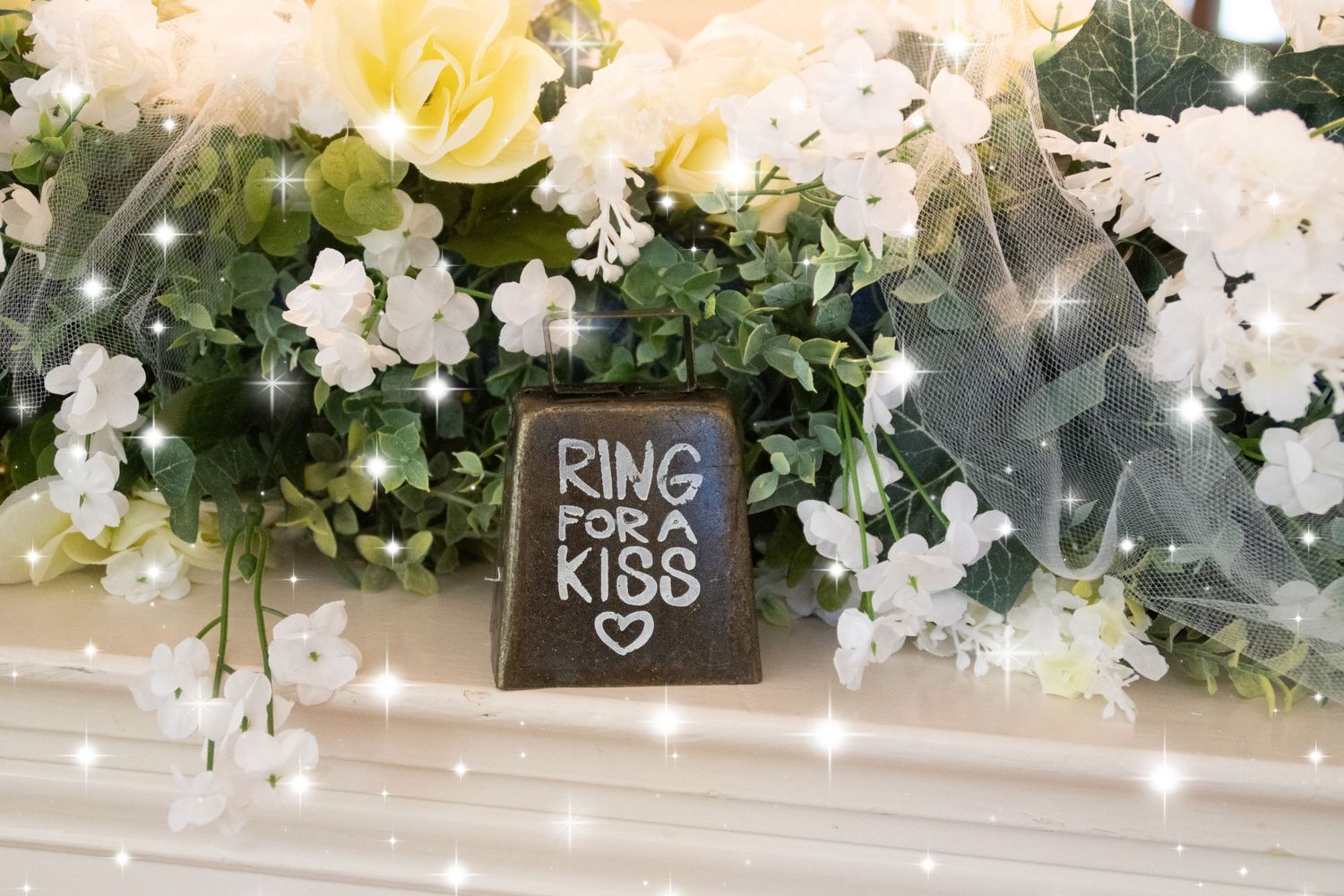 Small chalkboard sign reads 'Ring for a Kiss' surrounded by yellow and white flowers at a wedding.
