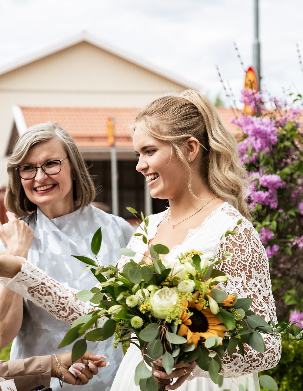 A wedding scene with a bride in lace holding a sunflower bouquet while sharing a joyful moment outdoors.
