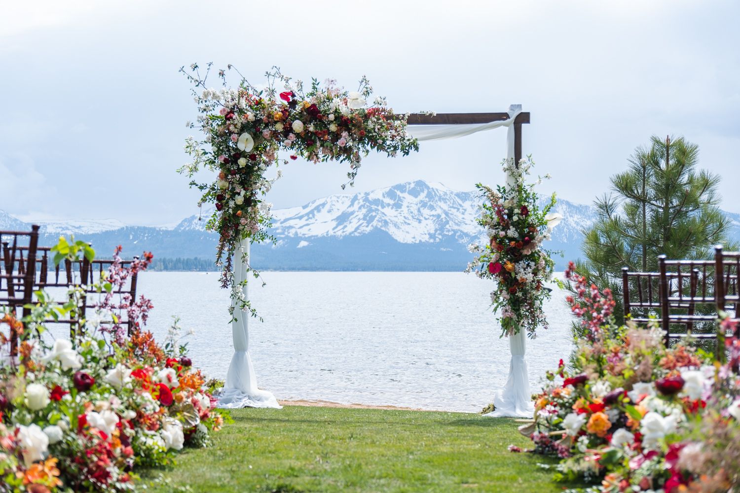 A white wedding arch adorned with pink and white flowers frames a stunning mountain lake view on a sunny day.