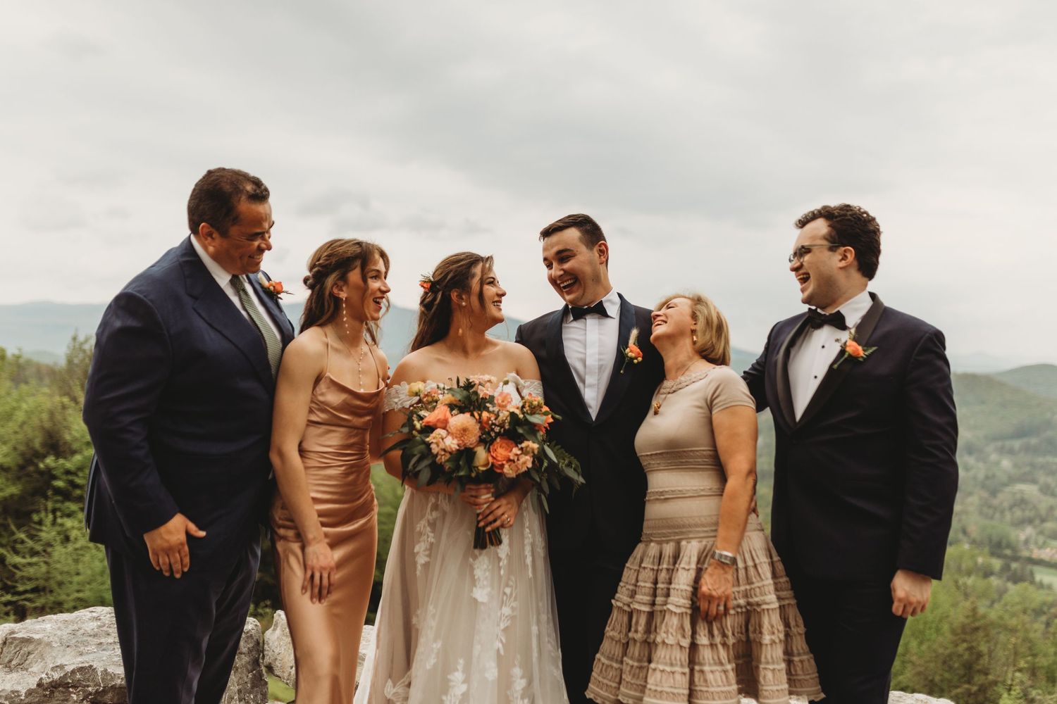 Wedding party in mix of formal attire poses together against scenic mountain backdrop with coral bouquets.