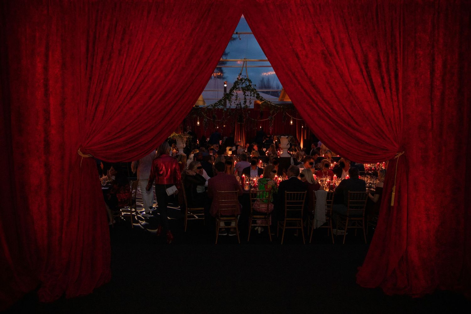 Red velvet curtains frame a dark dining room with elegant table settings and soft ambient lighting.