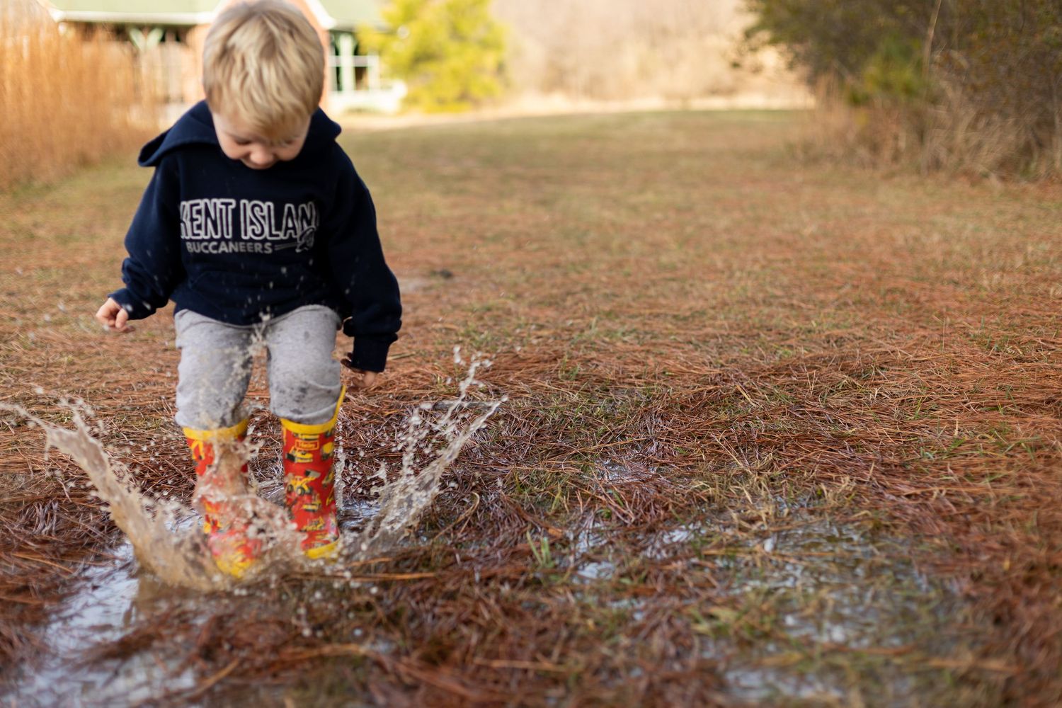 A child in rain boots makes a big splash in a puddle on a dirt path.