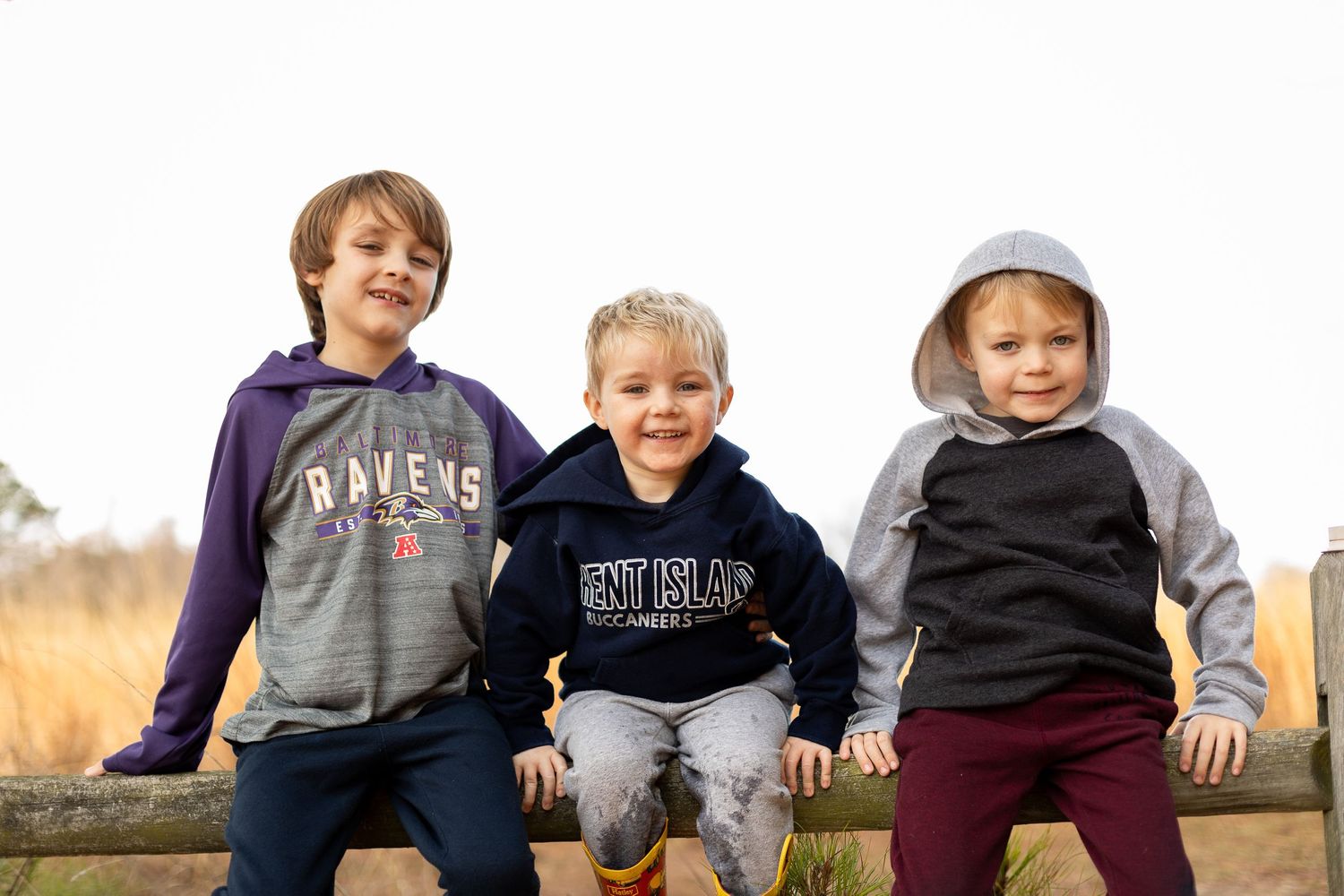 Three young children sitting together outdoors wearing casual sweatshirts and hoodies.
