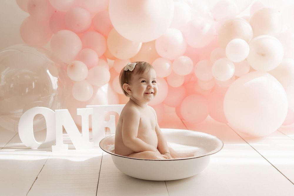 Baby sitting in white basin with pink balloon backdrop and 'ONE' text decoration.