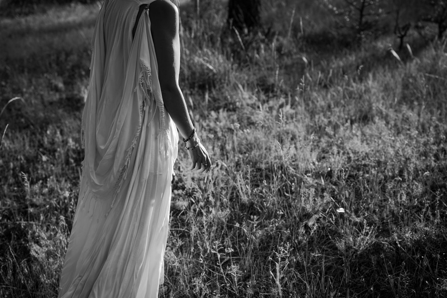 Figure in flowing white dress stands in a sunlit meadow of tall grass.