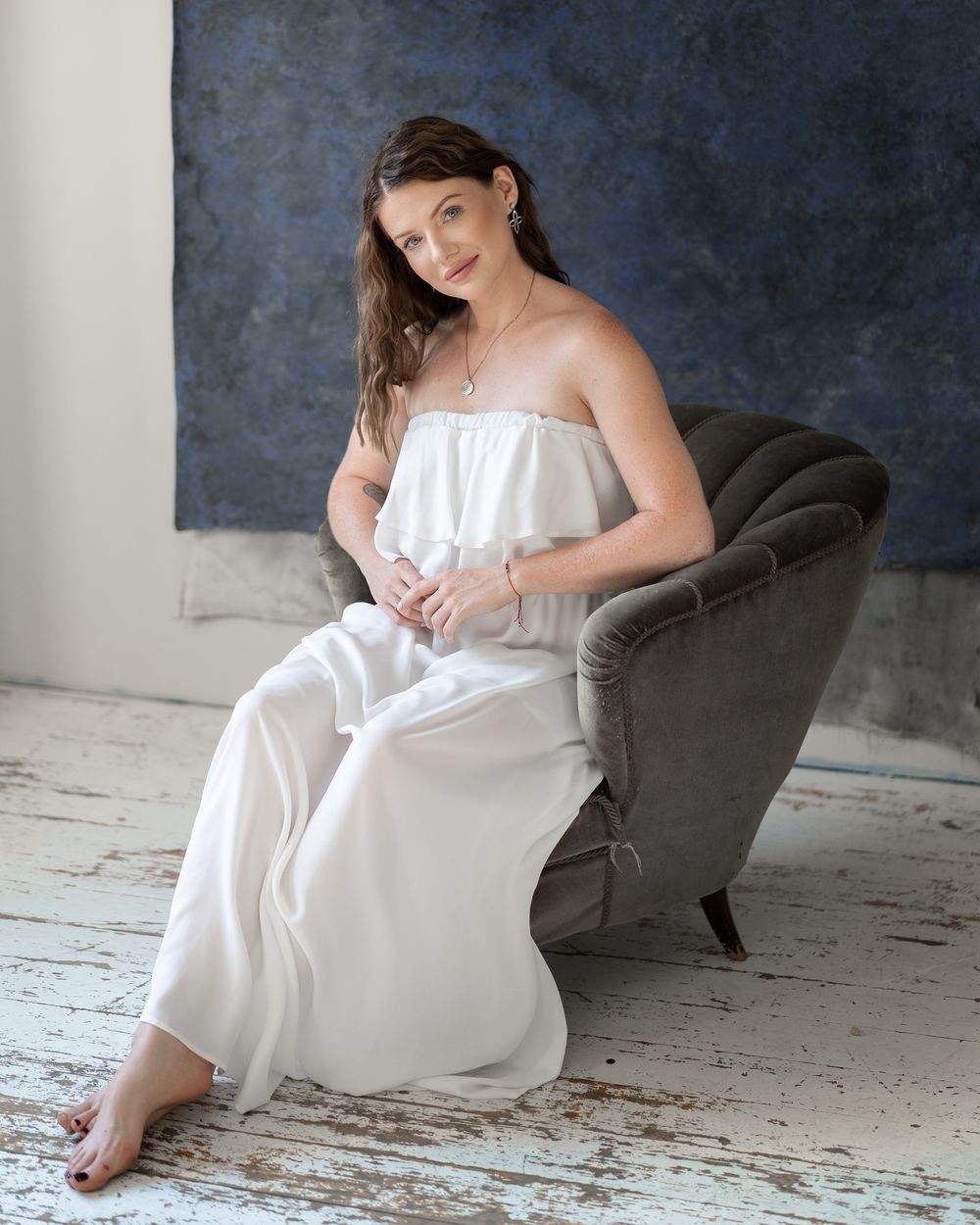 Person in white dress sits elegantly in grey velvet chair against dark blue wall.