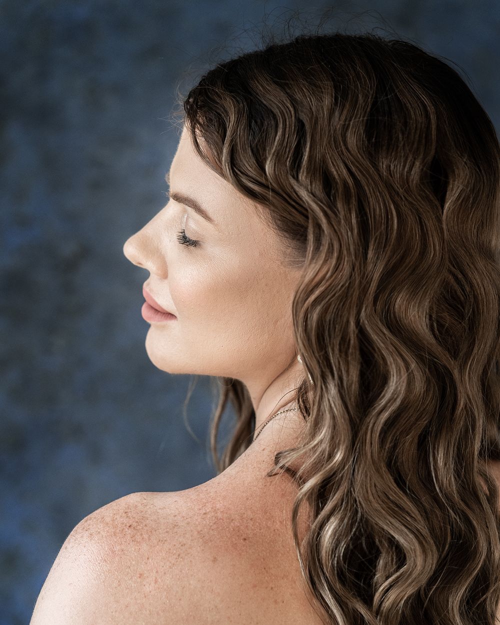 Side profile portrait showcasing wavy brown hair and natural makeup against dark backdrop.