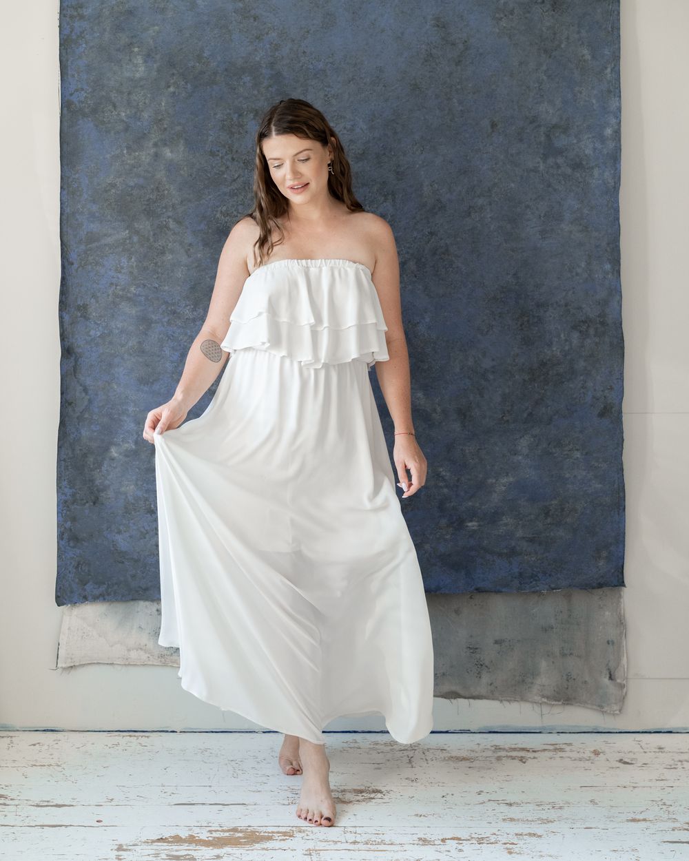 Woman in flowing white maxi dress dancing barefoot against blue studio backdrop.