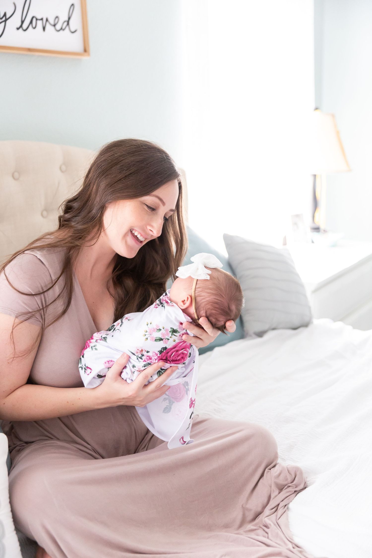 Mother in mauve dress holding sleeping baby in floral outfit on bed.