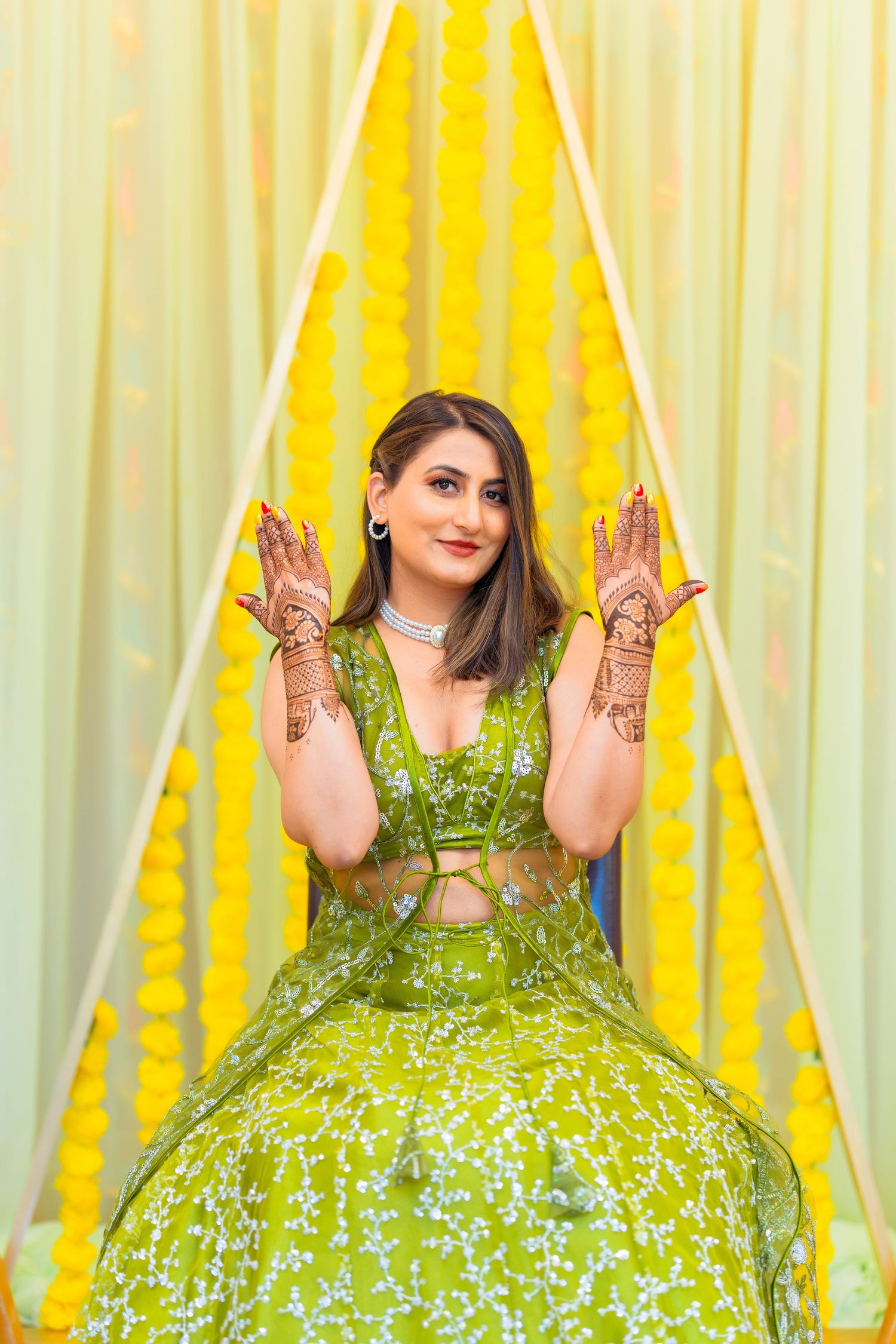 Woman in green lehenga shows intricate henna designs on her hands against yellow backdrop.