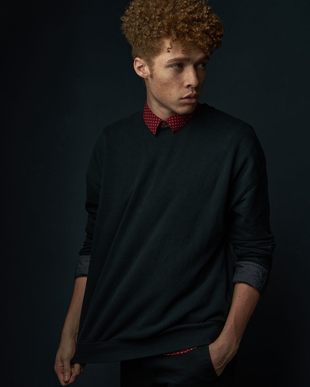 Young man with curly red hair wearing a dark green sweater and red collar shirt on dark background.