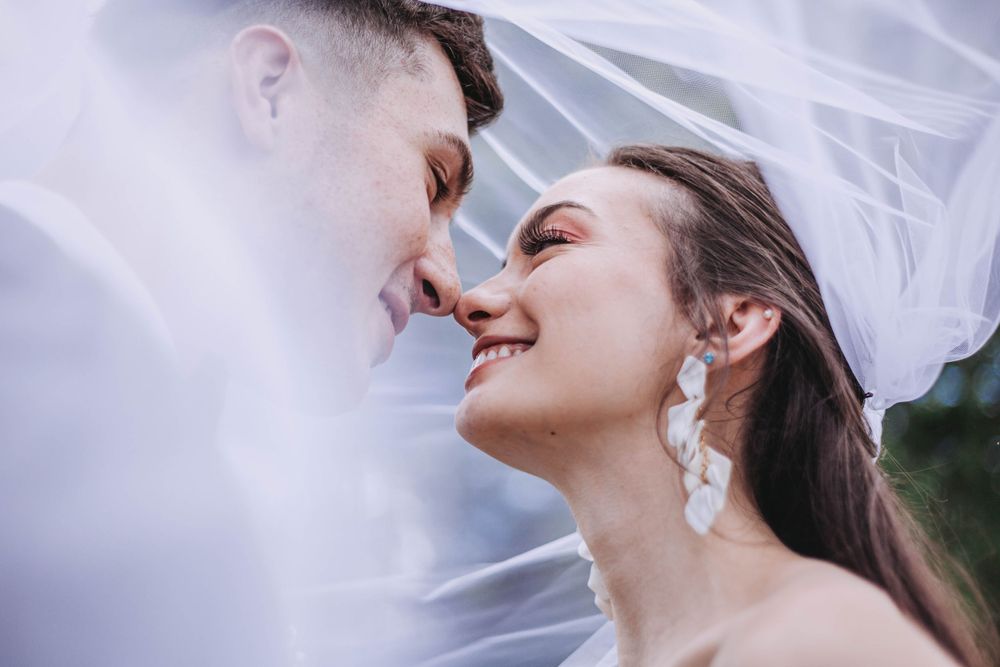 Bride and groom share a romantic moment under a flowing white veil, smiling close together.