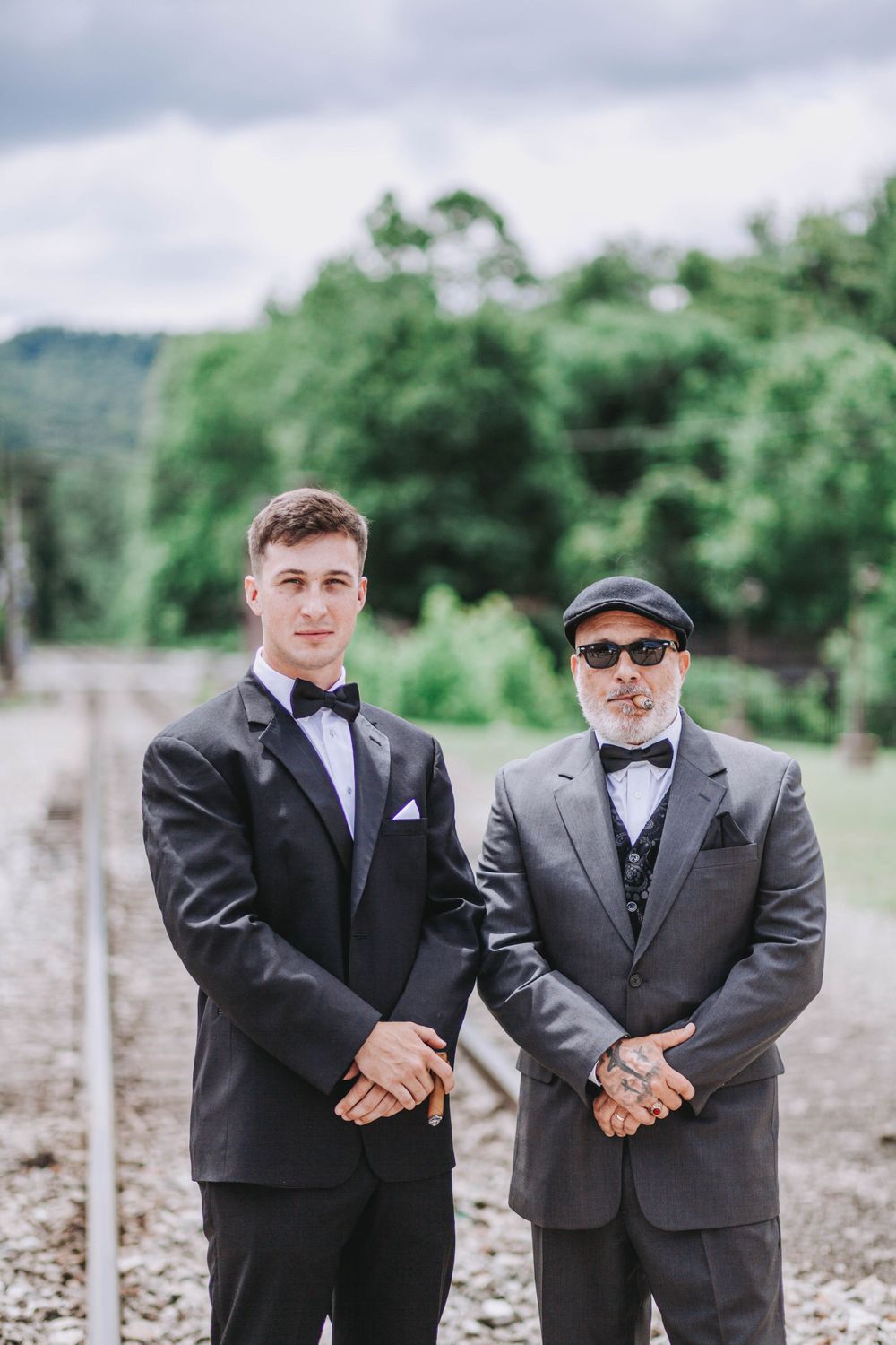 Groom and older gentleman in matching black tuxedos with bow ties posing near railway tracks.