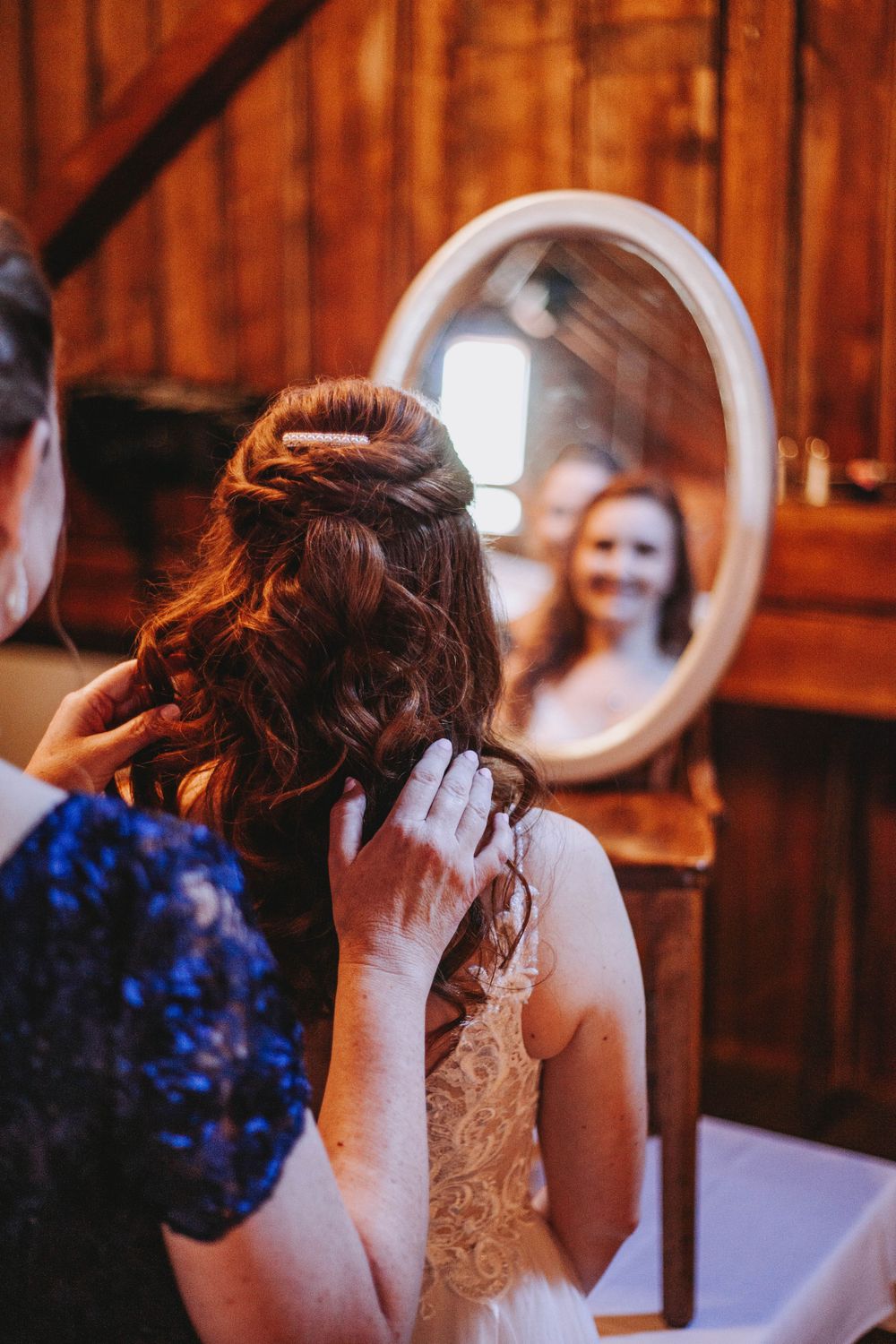 Hairstylist arranges bride's updo in a rustic wooden room as bride smiles in oval mirror.
