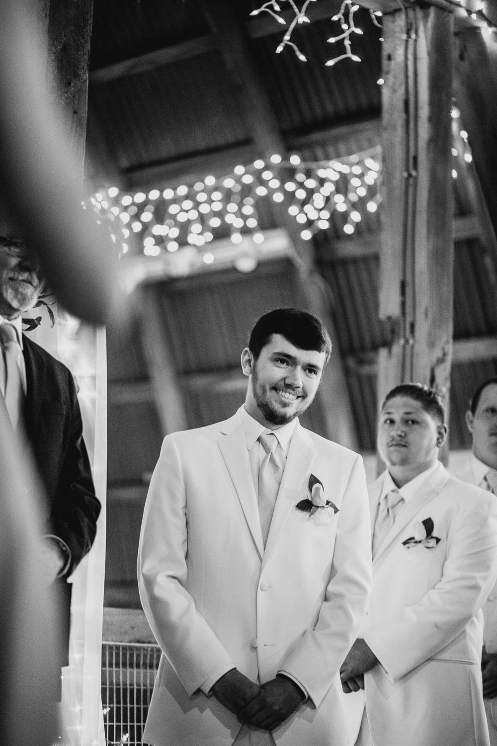 Groom in white suit smiling at the altar during rustic barn wedding ceremony with string lights.