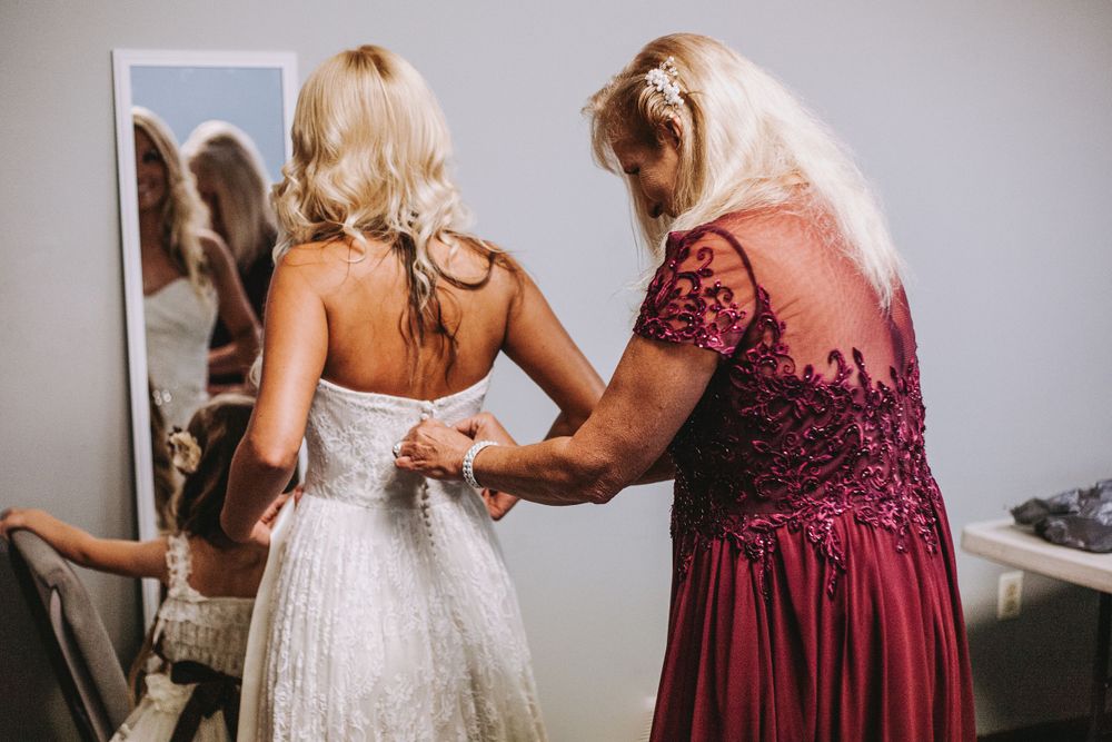 Mother in burgundy lace dress buttons up bride's white wedding gown before ceremony.