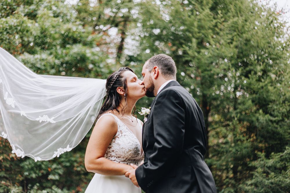 Bride and groom kissing outdoors as wedding veil flows in the wind among lush green trees.