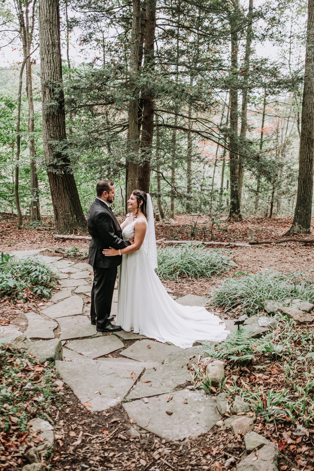 Bride and groom embrace on a stone garden path surrounded by tall trees in a wooded wedding setting.