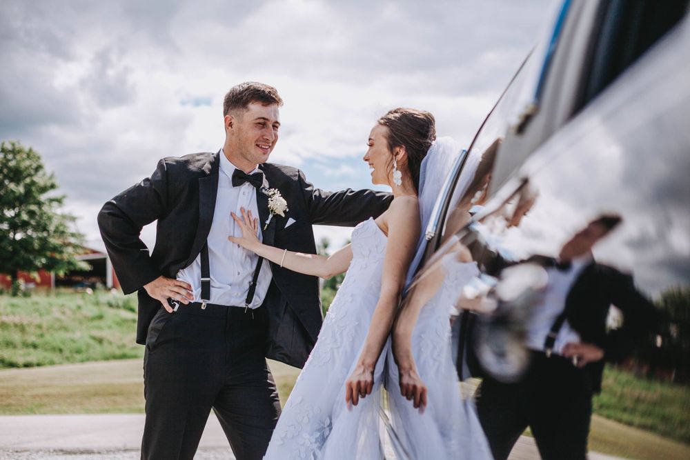 Laughing bride and groom share a playful moment beside a white limousine outdoors.