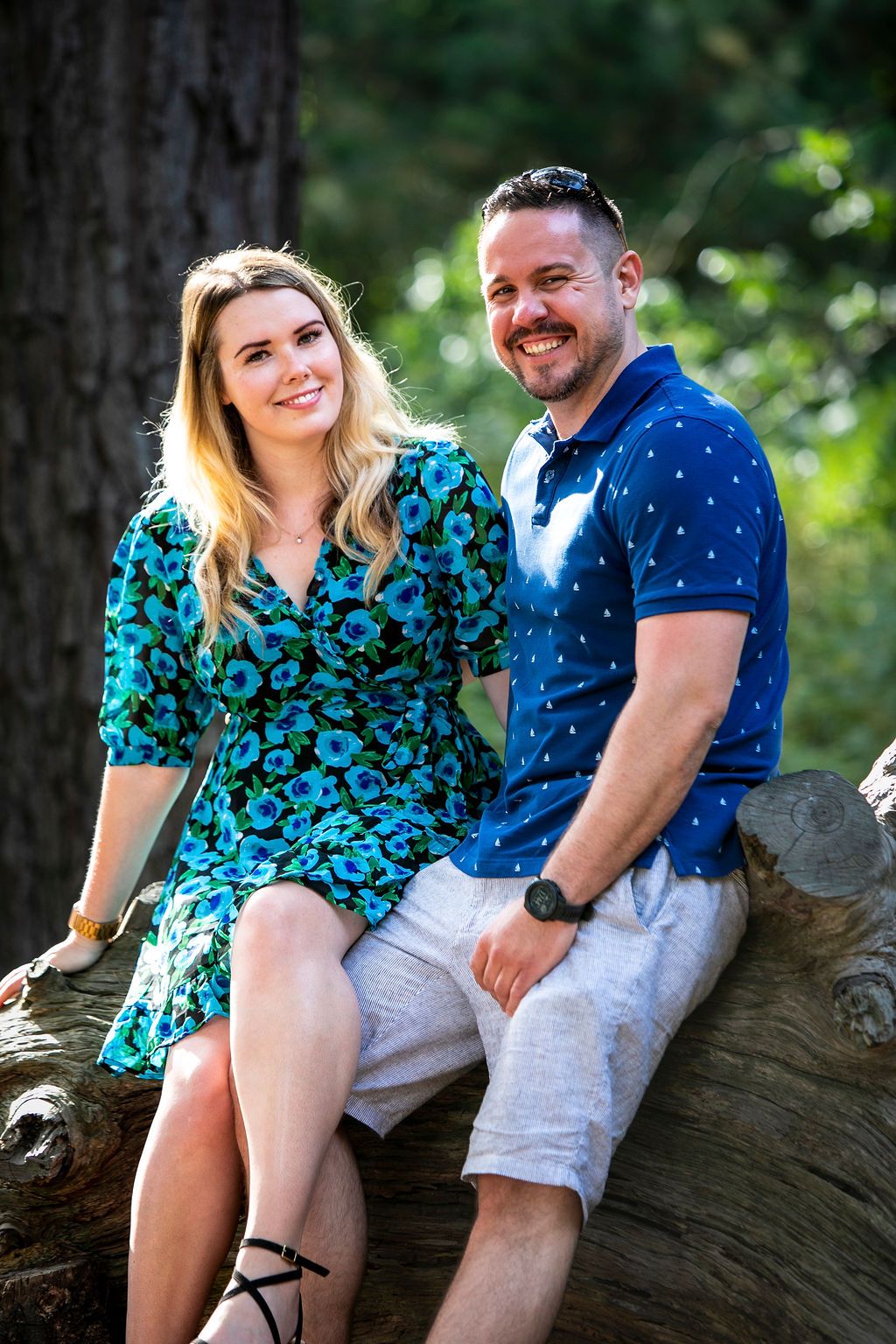 A happy couple sitting together on a fallen tree trunk in a sunny forest setting.