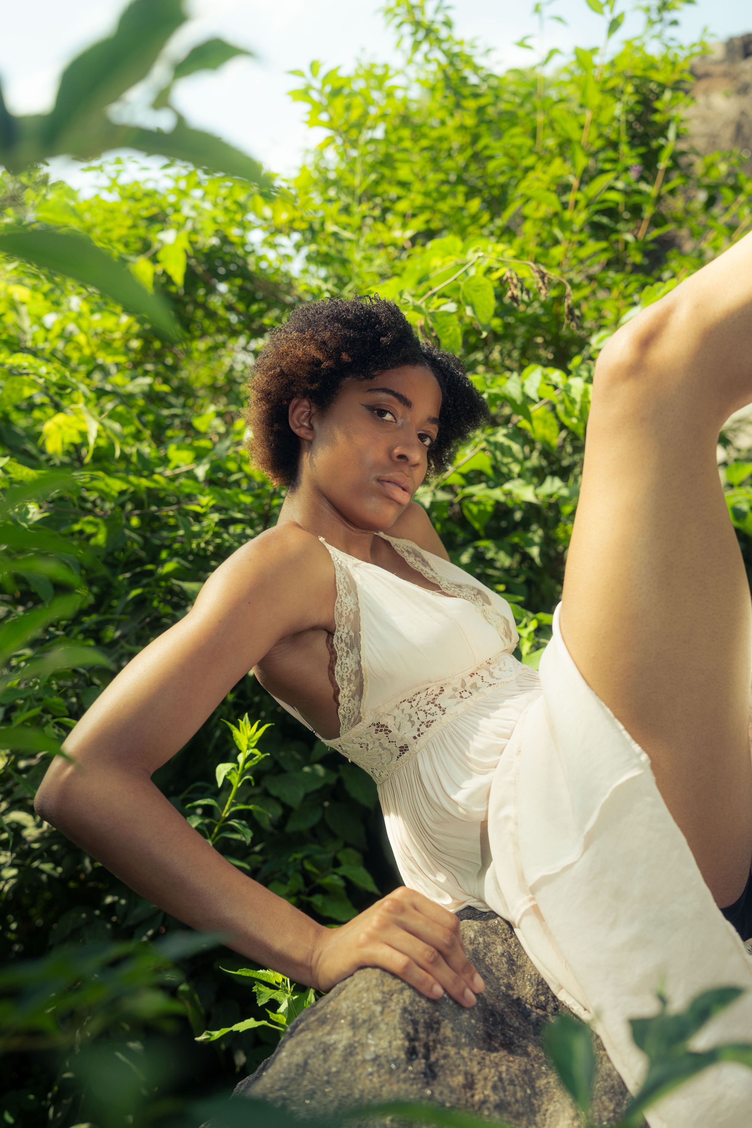 Figure in white lace dress poses gracefully against lush green foliage.
